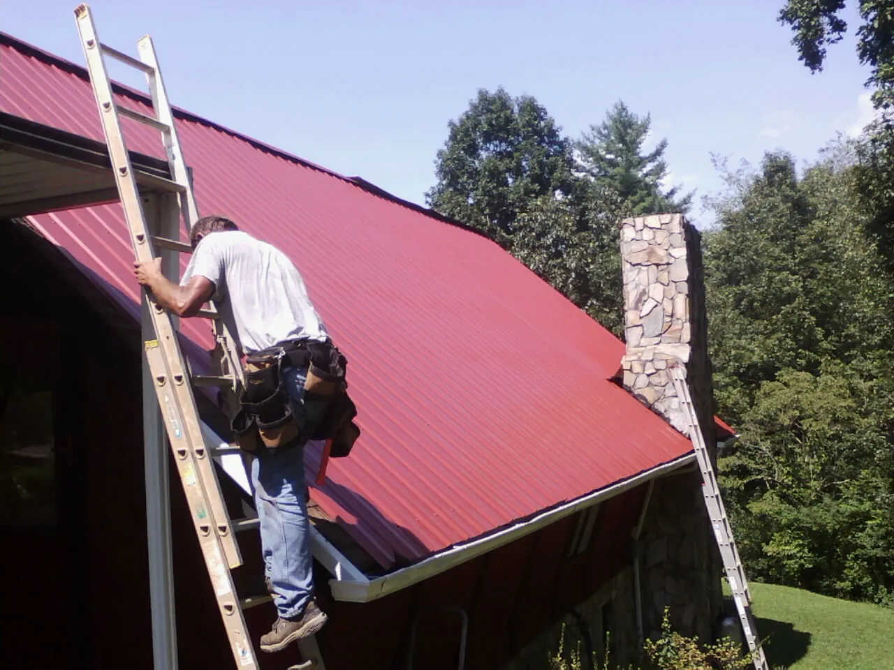 Roofer on a ladder beside a red metal roof and stone chimney — Soffit & Fascia Repair by Roofing Brothers in Rollinsford